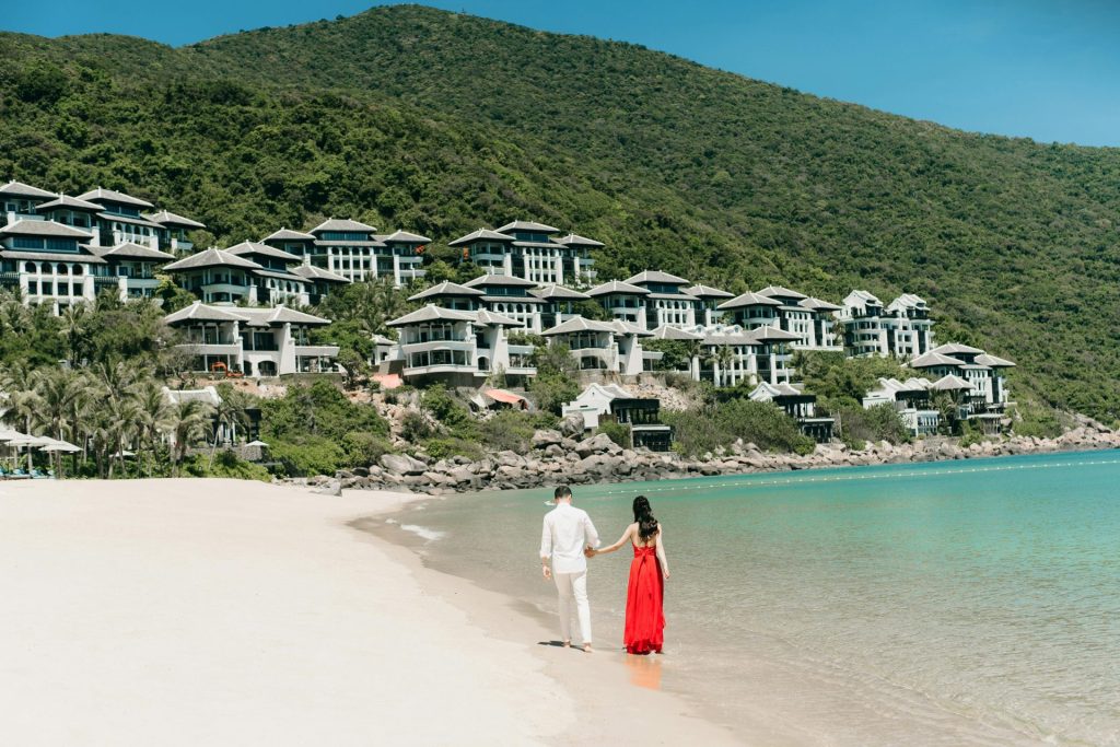 A couple walks hand in hand along a scenic beach next to luxurious villas, enjoying a sunny day.