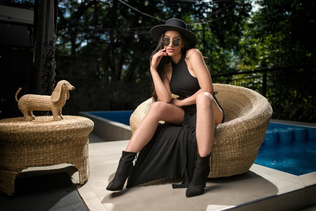 Stylish woman in black dress and hat sitting by a pool, exuding luxury and elegance.